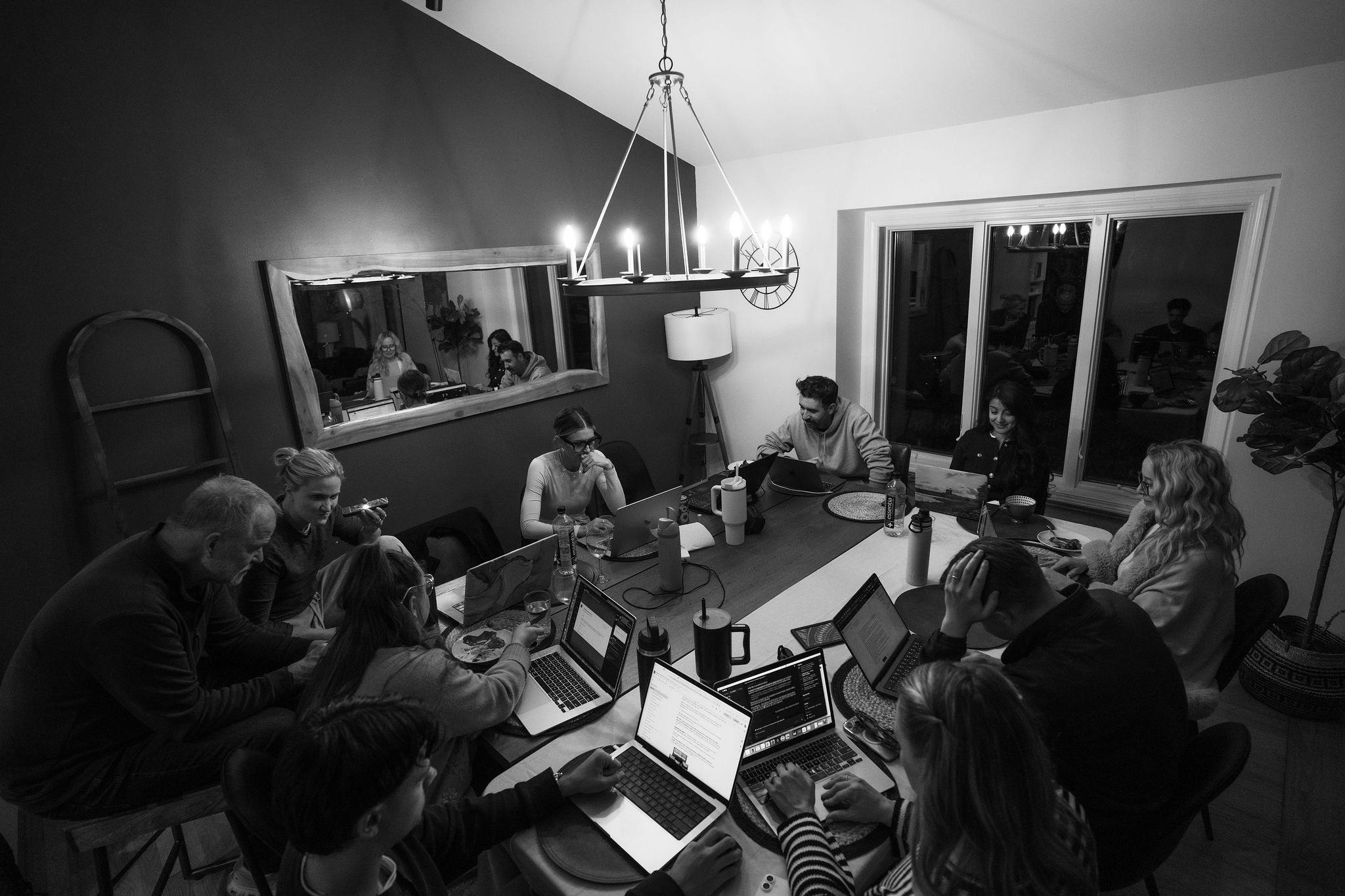 The whole crew working late around the dining table, laptops open under the chandelier